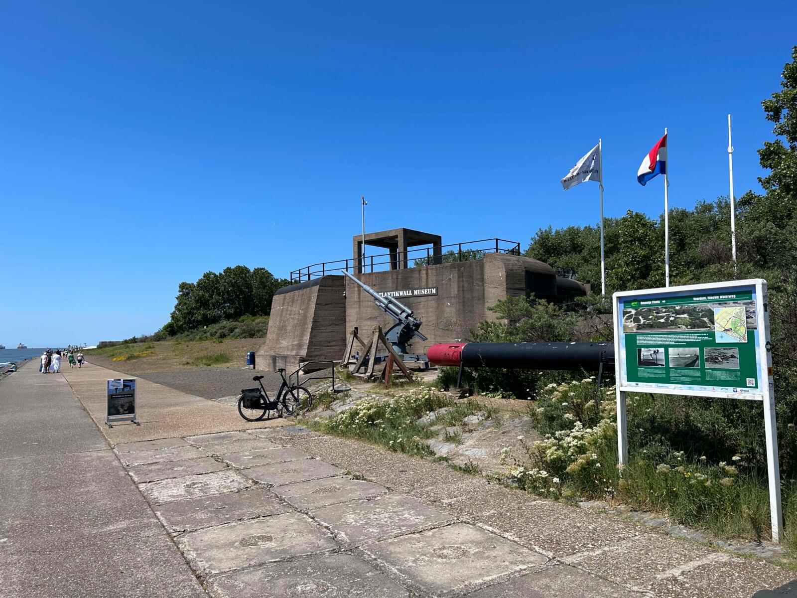 Een historische bunker van het Atlantikwall Museum op een zonnige dag, met een stuk militair geschut buiten, een informatiebord, wandelpad en de Nederlandse vlag in de duinen.