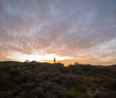 Het silhouet van een wandelaar op een duin of bunker van de Atlantikwall, met een dramatische, kleurrijke zonsondergang met oranje en paarse wolken op de achtergrond.