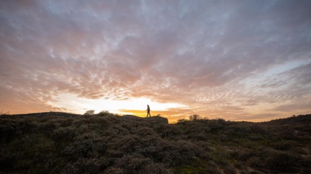 Het silhouet van een wandelaar op een duin of bunker van de Atlantikwall, met een dramatische, kleurrijke zonsondergang met oranje en paarse wolken op de achtergrond.