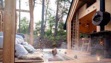 Een persoon geniet van een stomende hottub verzonken in het houten terras van een moderne cabin in het bos, met uitzicht op hoge bomen en een knusse houtkachel ernaast.
