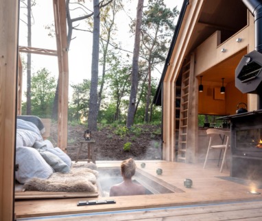 Een persoon geniet van een stomende hottub verzonken in het houten terras van een moderne cabin in het bos, met uitzicht op hoge bomen en een knusse houtkachel ernaast.