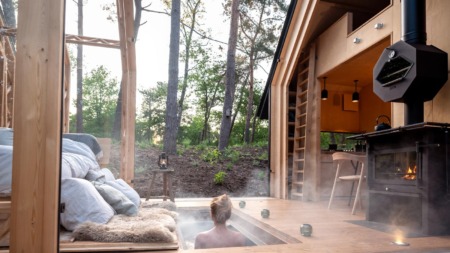 Een persoon geniet van een stomende hottub verzonken in het houten terras van een moderne cabin in het bos, met uitzicht op hoge bomen en een knusse houtkachel ernaast.