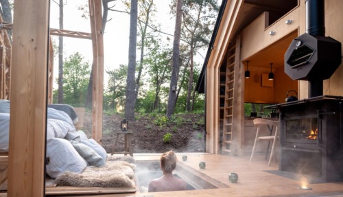 Een persoon geniet van een stomende hottub verzonken in het houten terras van een moderne cabin in het bos, met uitzicht op hoge bomen en een knusse houtkachel ernaast.
