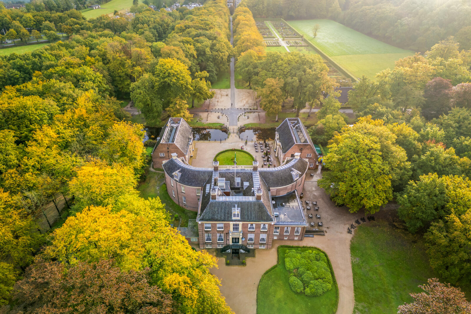 Een majestueuze luchtfoto van Kasteel Groeneveld in Baarn, omgeven door een uitgestrekt landgoed met bomen in volle herfstkleuren, een formele tuin en een lange oprijlaan.