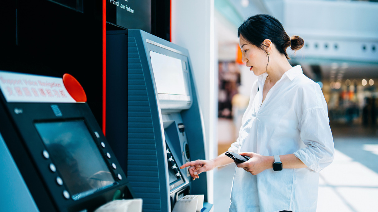 Jonge Aziatische vrouw die een geldautomaat (ATM) gebruikt in een winkelcentrum. Geld opnemen, rekeningen betalen, het saldo controleren en een overschrijving doen. Persoonlijk bankieren. Bescherming van de privacy. Beveiliging van mobiel bankieren. Zelfbedieningsconcept