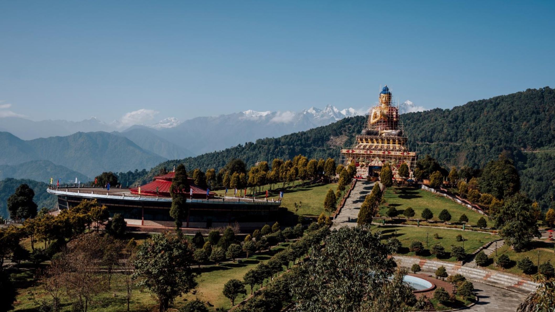 Een panoramisch uitzicht over het Buddha Park van Ravangla in Sikkim, met een enorm gouden Boeddhabeeld rechts, een rond gebouw met een rood dak links, en op de achtergrond de besneeuwde toppen van de Himalaya.