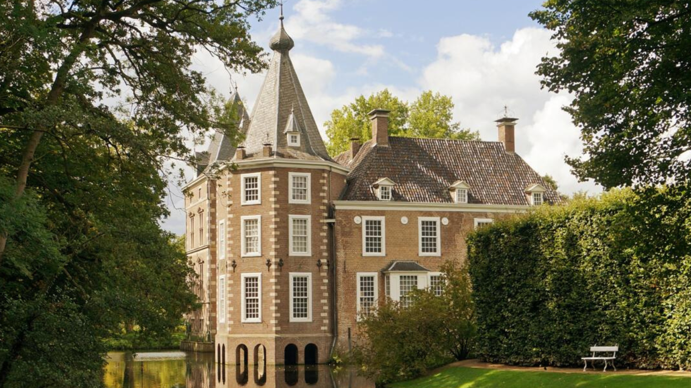 Een schilderachtig uitzicht op het Kasteel. met zijn karakteristieke toren en bakstenen gevel, weerspiegeld in het omringende water en omgeven door weelderige groene bomen en een hoge haag.