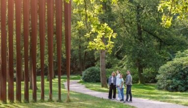 Vier personen die op een bospad in het beeldenpark van het Kröller-Müller Museum in Gelderland omhoog kijken naar het grote cortenstalen kunstwerk Palissade van Evert Strobos.