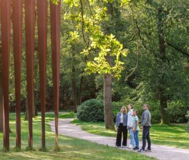 Vier personen die op een bospad in het beeldenpark van het Kröller-Müller Museum in Gelderland omhoog kijken naar het grote cortenstalen kunstwerk Palissade van Evert Strobos.