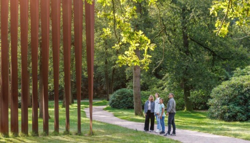Vier personen die op een bospad in het beeldenpark van het Kröller-Müller Museum in Gelderland omhoog kijken naar het grote cortenstalen kunstwerk Palissade van Evert Strobos.