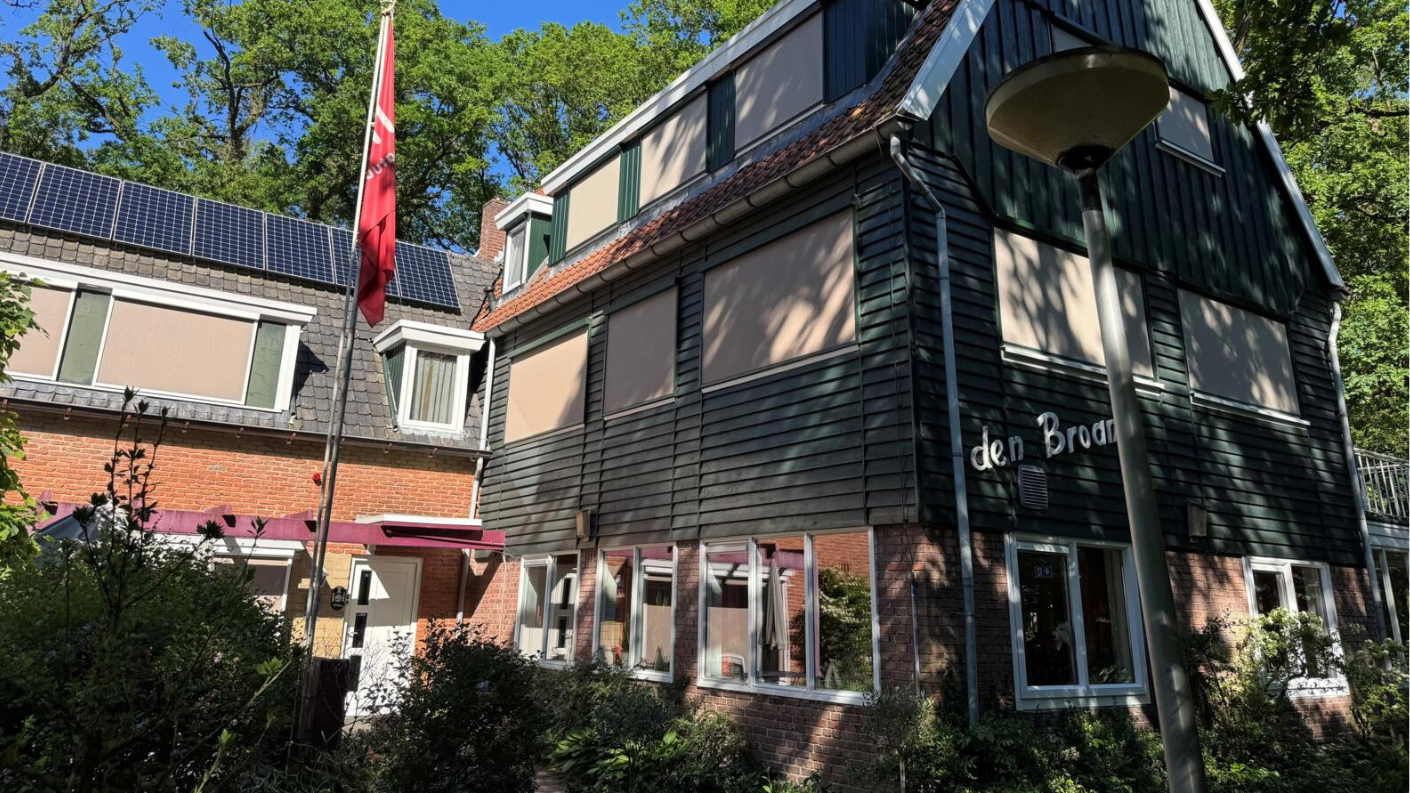 De gevel van Hotel Den Broam in Eibergen, een combinatie van een traditionele boerderij met donkergroene houten gevelbekleding en een moderner deel met zonnepanelen, omgeven door groen.