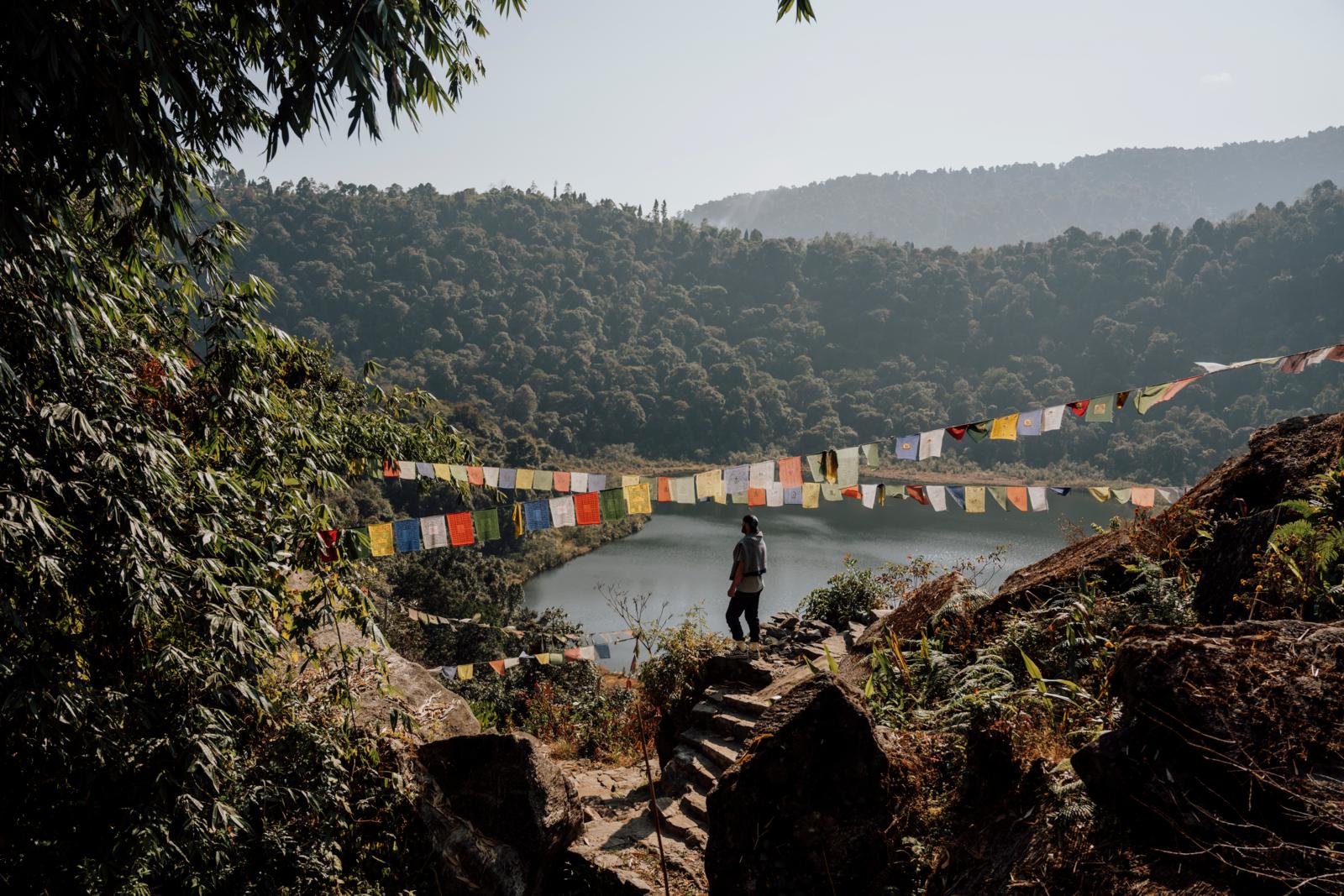 Een persoon staat op een rotsachtig pad met een trap, uitkijkend over het serene Khecheopalri-meer in Sikkim, omgeven door dichte groene bossen en kleurrijke boeddhistische gebedsvlaggen die over het water hangen.