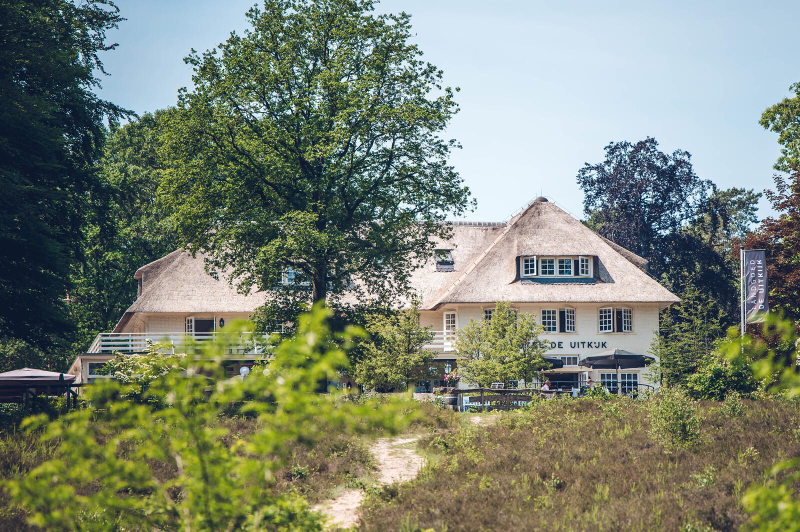 Het statige landhuis van Landgoed De Uitkijk in Hellendoorn, met zijn karakteristieke rieten dak en witte gevel, gedeeltelijk verscholen achter groene struiken en bomen, met een zandpad op de voorgrond.