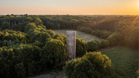Een moderne uitkijktoren reikt hoog boven de boomtoppen uit in een groen bosgebied tijdens de zonsondergang.