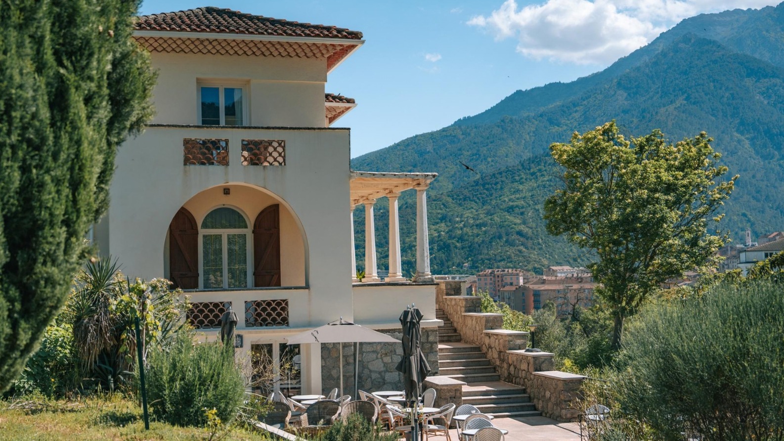 Exterieur van Hôtel Si Mea met terras, stenen trap en bergen op de achtergrond op Corsica