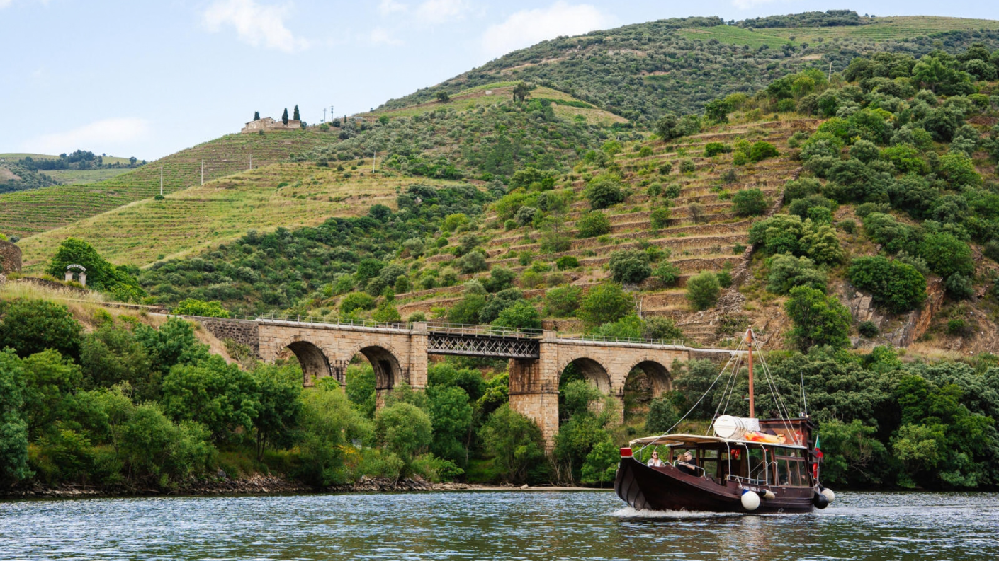 Een traditionele rabelo-boot op de Douro-rivier met een stenen spoorbrug en steile wijngaardterrassen op de achtergrond