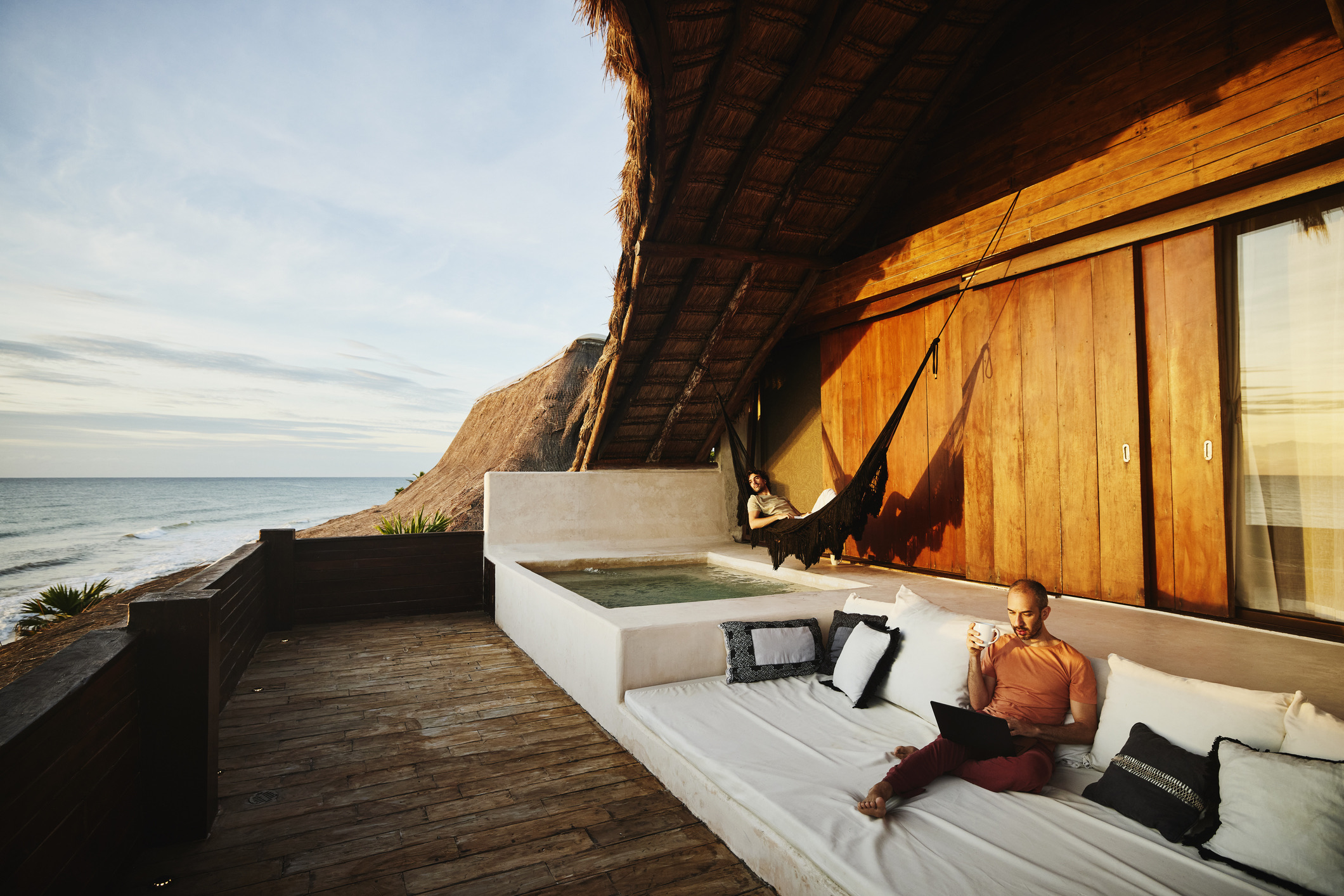Breed shot van een man die koffie drinkt en op zijn laptop werkt, terwijl zijn partner bij zonsopgang in een hangmat op het terras van een luxe tropische strandvilla ligt te ontspannen