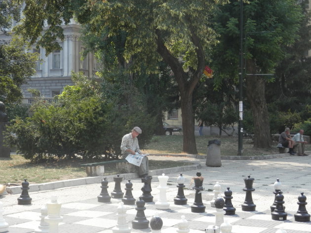 Oud mannetje zit op een bankje een krant te lezen voor een groot schaakspel, in een park in Sarajevo. 