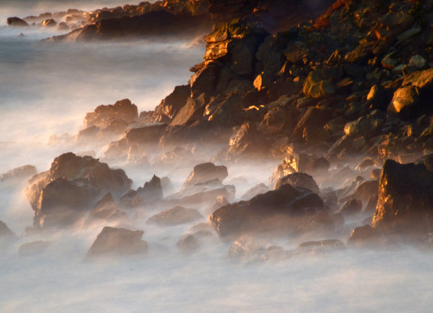 Strandimpressie met verneveld zeewater en laat licht op rotsen.